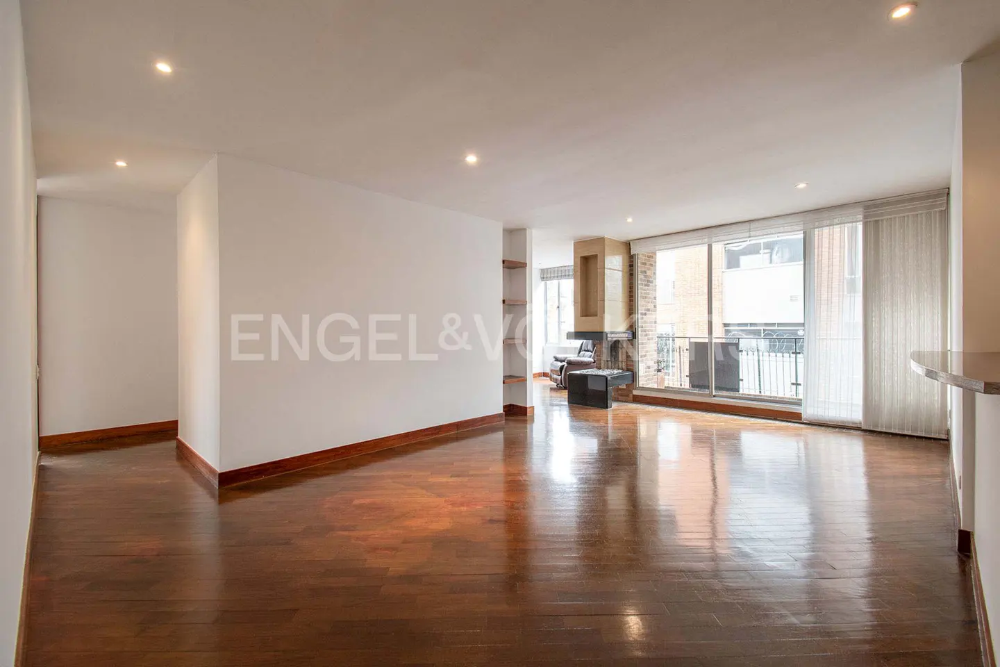 Bright, empty living room with hardwood floors, white walls, and a fireplace. Large windows offer city views. Engel & Völkers logo on the wall.