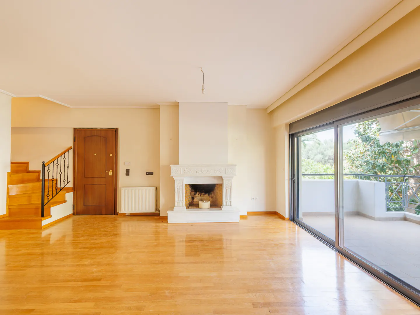 Bright, empty living room with wood floors, stairs, and a white fireplace. Sliding glass doors lead to a balcony with trees visible.