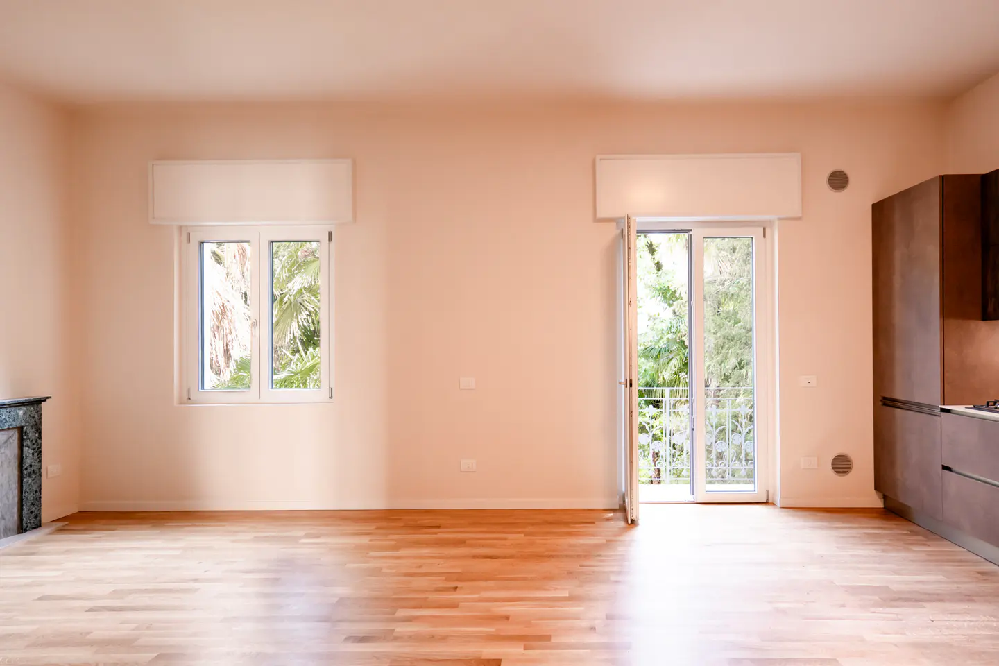 Empty room with light wood floors, beige walls, a window, a balcony door, and a modern dark kitchen cabinet unit.