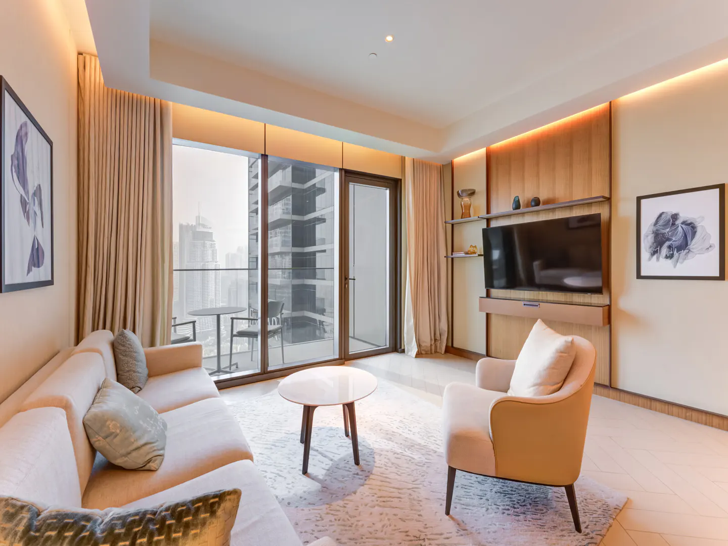 Living room with beige sofa, chair, and round table. Balcony with city view. TV on wood panel. Artwork on walls.