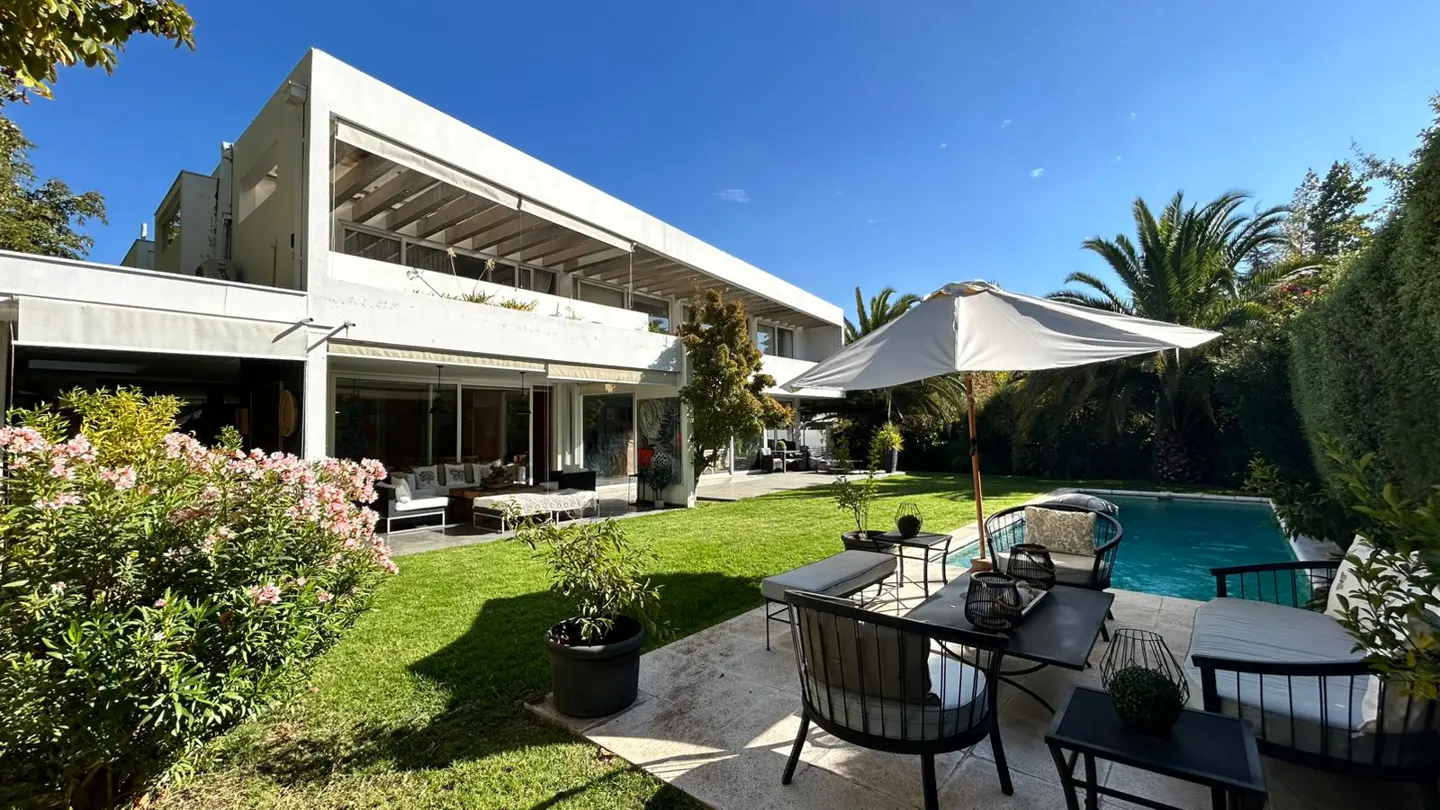 Backyard view of a modern white house with a pool, patio furniture, green lawn, and lush greenery under a clear blue sky.