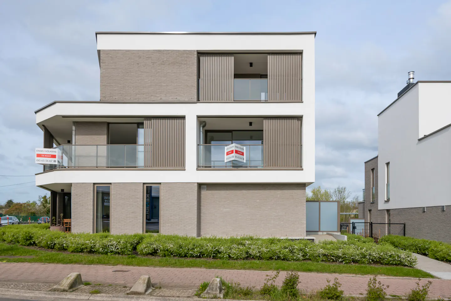 Modern three-story apartment building with balconies and "For Sale" signs. The building is white and gray brick with vertical slat blinds.