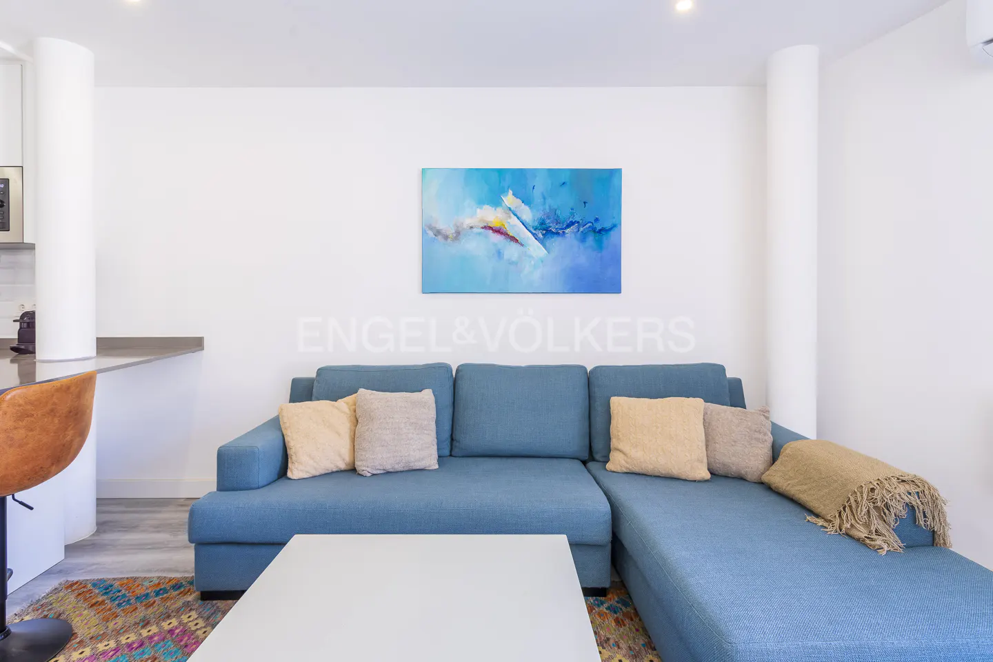 Living room with a blue sectional sofa, pillows, and a white coffee table. An abstract blue painting hangs above the sofa.