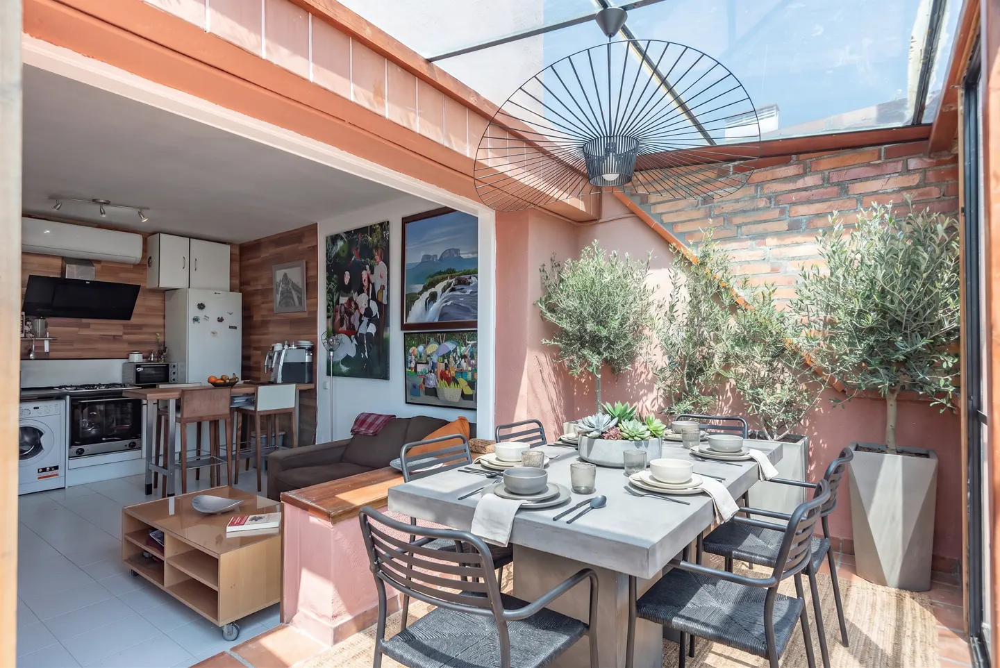 Bright, open-plan living space with a dining area under a glass roof, featuring a modern table setting and potted olive trees.