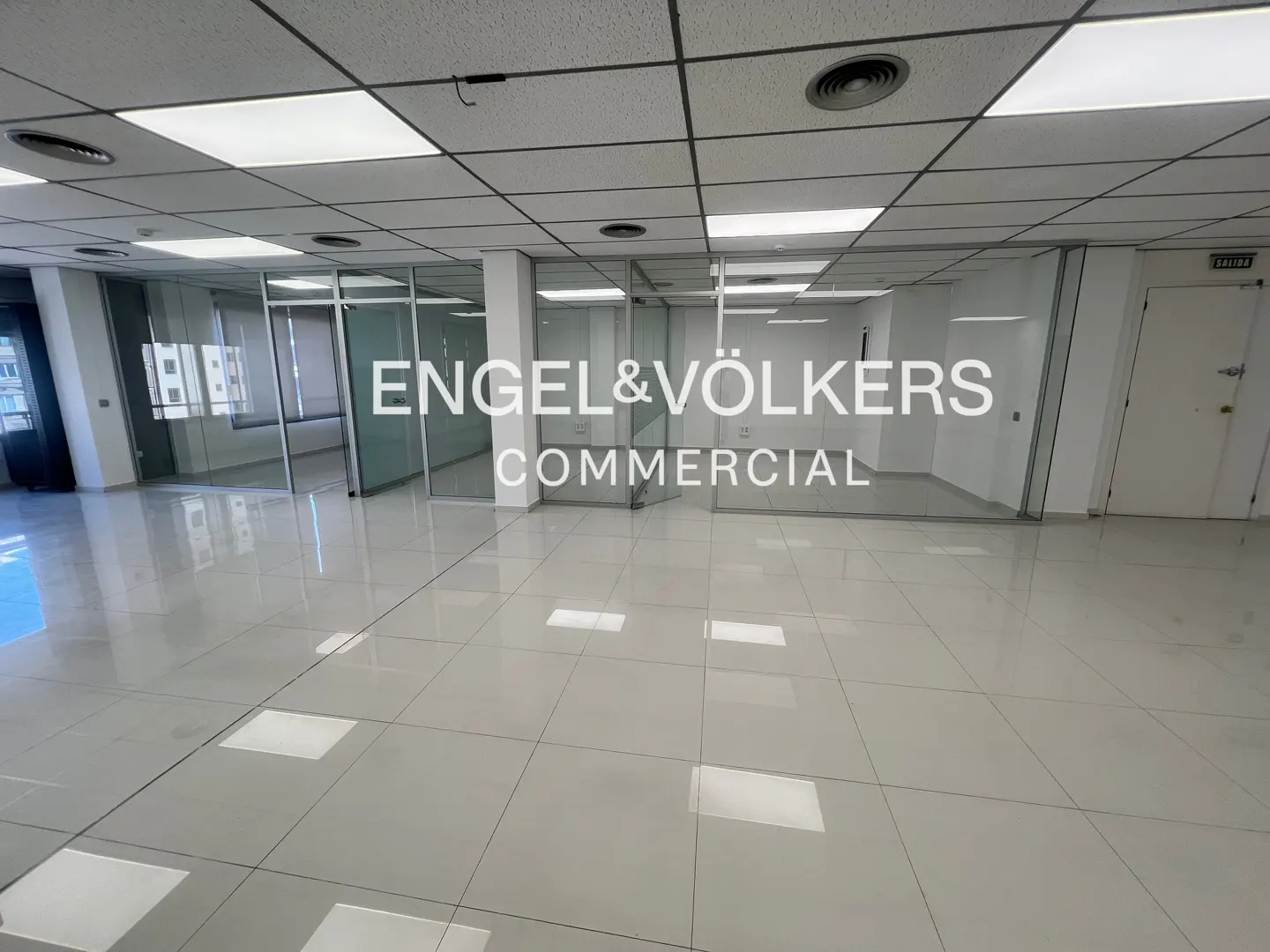 Empty office space with glass-walled rooms, white tile floor, and "Engel & Völkers Commercial" logo on the glass.