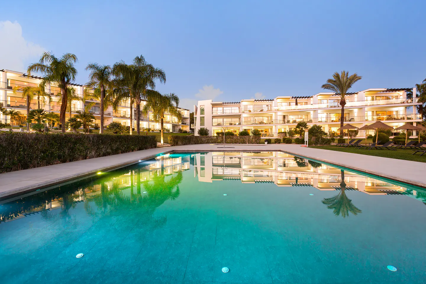 Turquoise pool reflects white condos with balconies and palm trees under a blue sky at dusk.