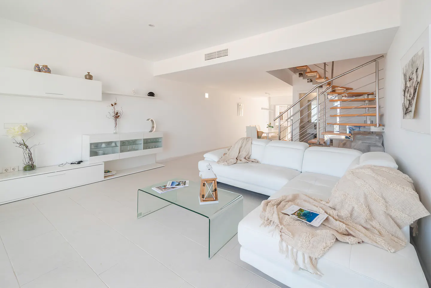 Bright, modern living room with white walls, floors, and furniture. A glass coffee table sits in front of a white sectional sofa with throws. Stairs are visible in the background.