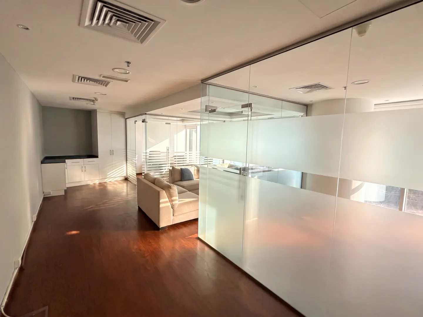 An office space with a tan sofa behind frosted glass walls on a dark wood floor.