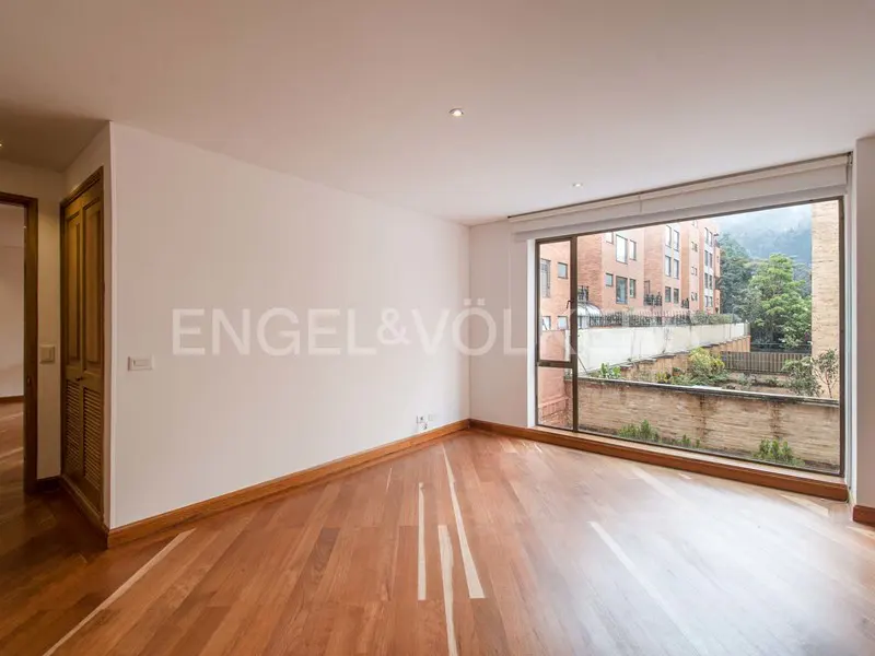 Empty room with hardwood floors, white walls, and a large window showing a brick building and greenery.