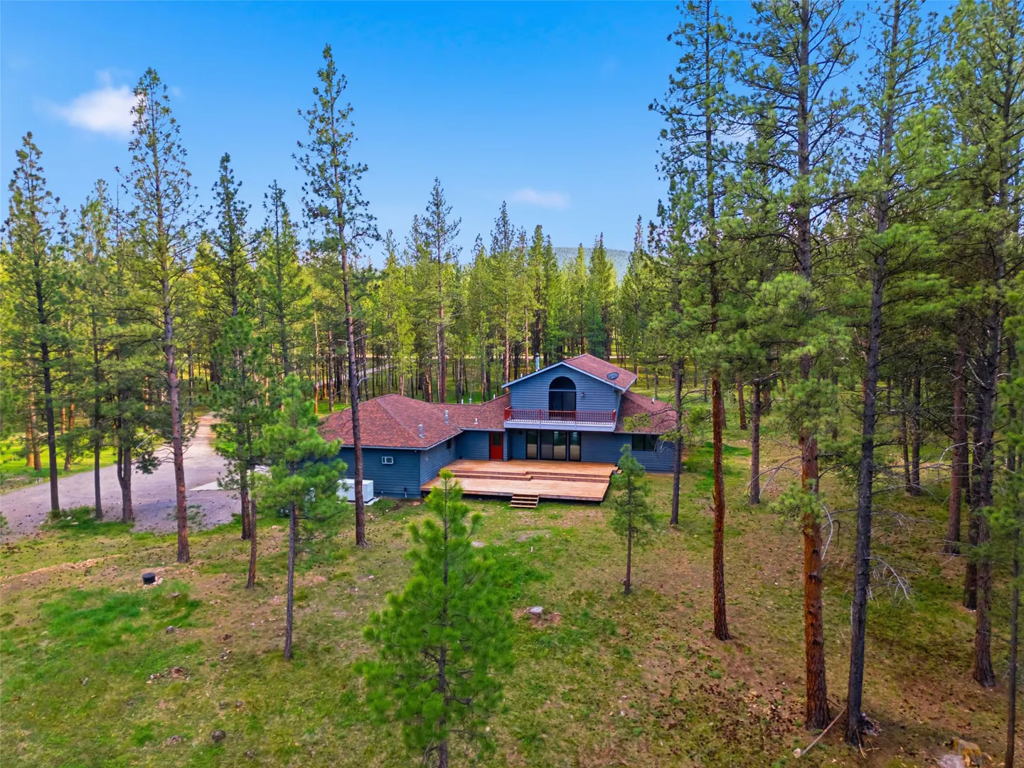 Two-story blue house with a red roof and deck surrounded by tall green trees under a blue sky.