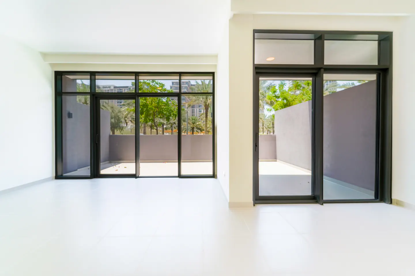 Bright, empty room with white walls and floor. Black-framed sliding glass doors lead to outdoor patios with trees and greenery.
