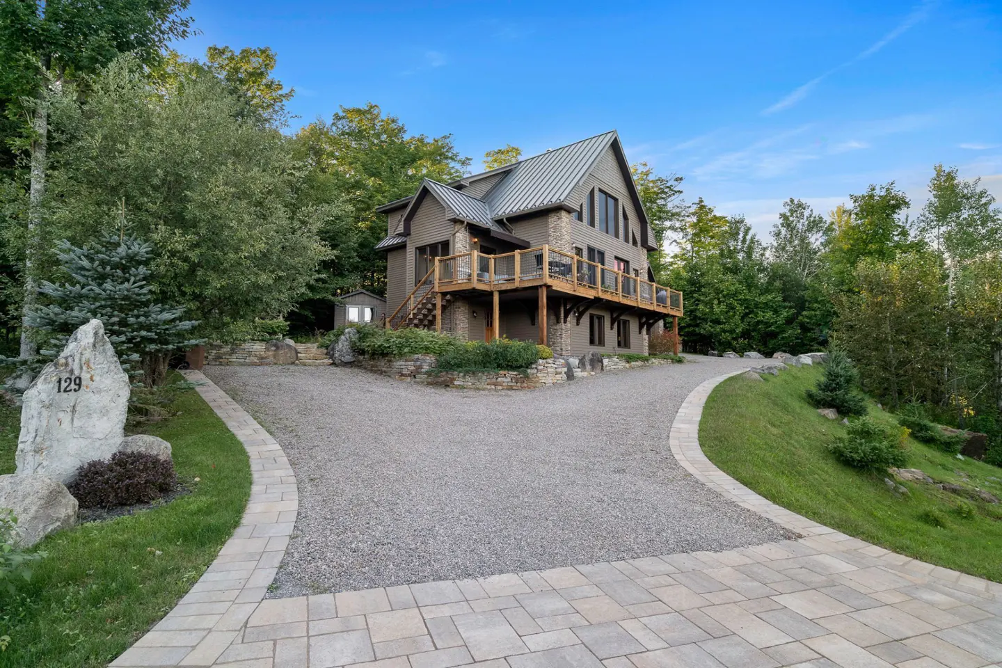 Two-story house with a metal roof, a wooden deck, and a gravel driveway surrounded by trees. A stone with the number 129 is on the left.