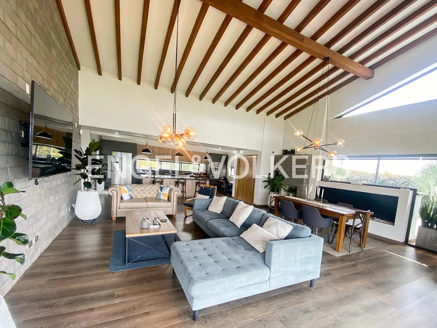 Open-concept living room with blue sofa, wood floors, and exposed beam ceiling. Dining table and kitchen visible in background.