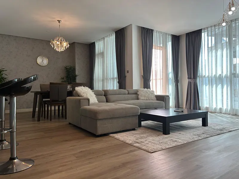 Living room with a gray sectional sofa, black coffee table, and dining area with a chandelier. Curtains frame large windows.