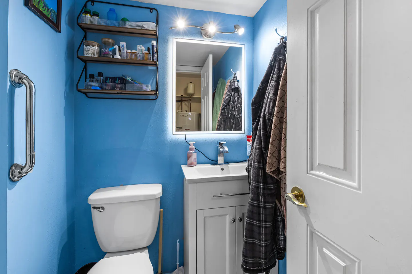 Small blue bathroom with a white toilet, vanity, illuminated mirror, wall shelving, and robes hanging on the door.