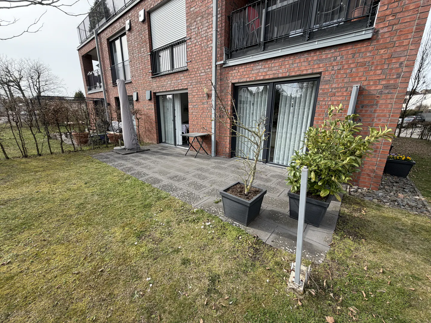 Brick apartment building with a patio, lawn, and hedge. Potted plants sit on the patio. Balconies are visible on the upper floors.