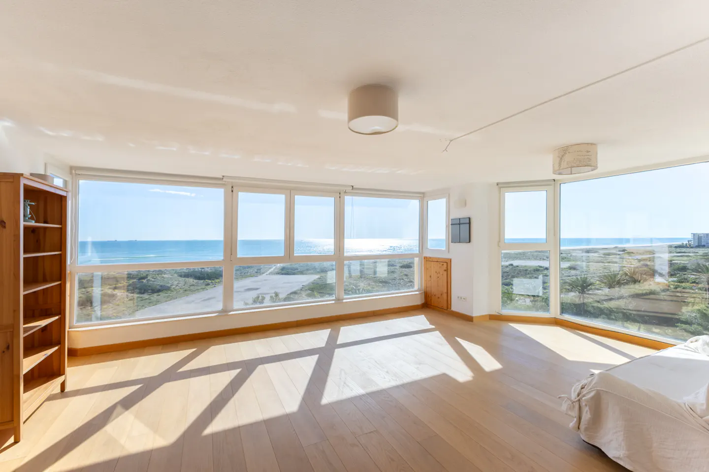 Bright, airy room with wood floors, large windows overlooking the ocean, and a wooden bookcase. Sunlight streams in.