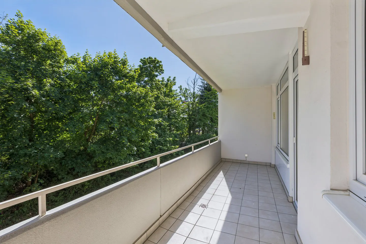 A long, covered balcony with light-colored tiled flooring, a metal railing, and a view of lush green trees under a blue sky.