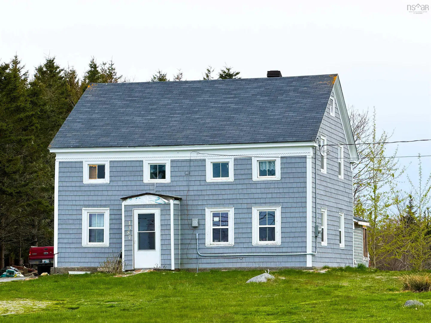 Two-story gray house with white trim and a gray shingled roof on a green lawn. Trees in the background.