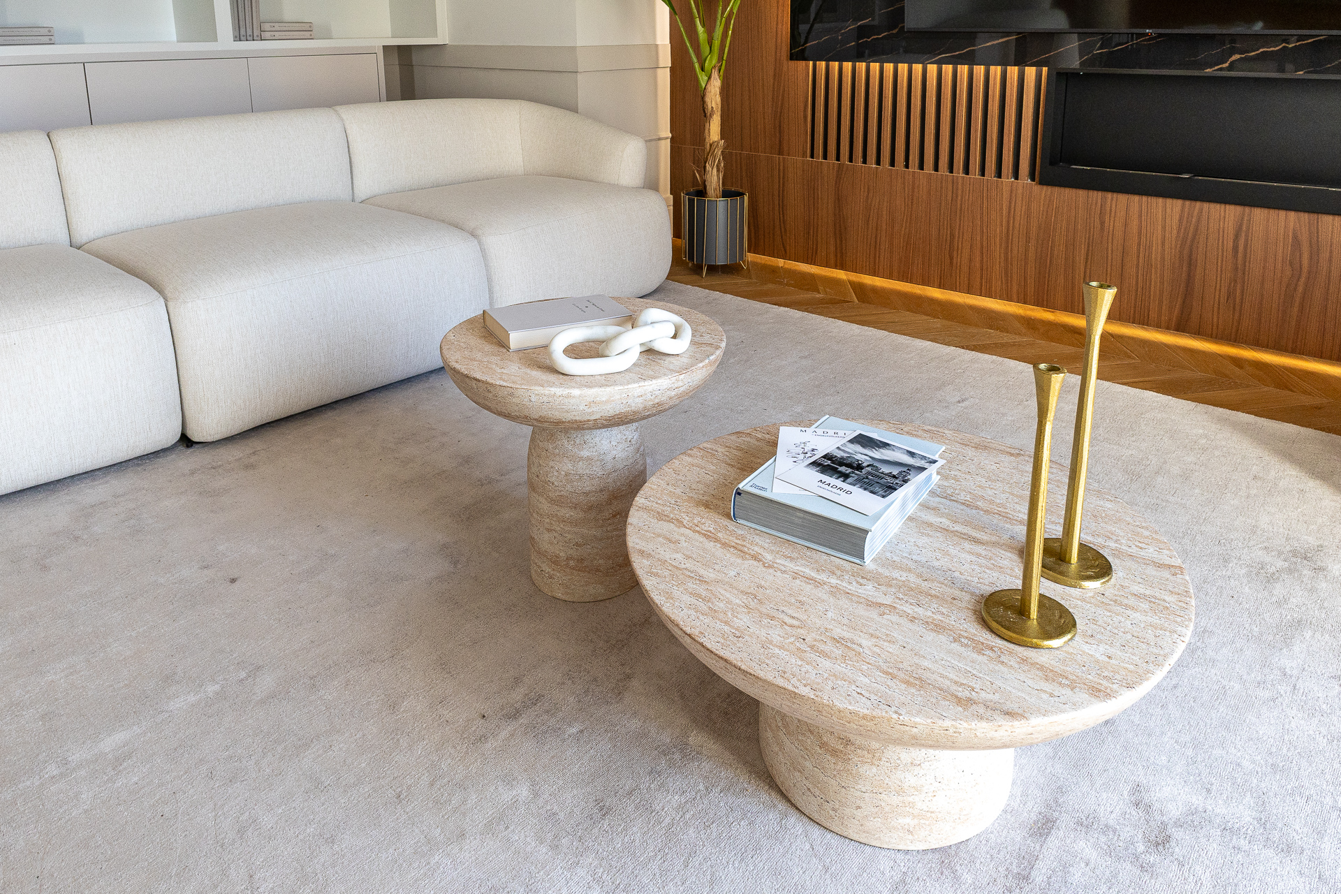 Modern living room with a white sofa, two round travertine coffee tables, gold candlesticks, and a wood-paneled wall. [AI-generated text]