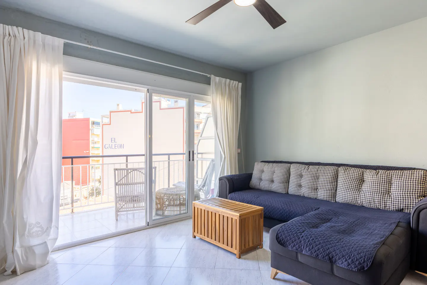 A bright living room with a blue sofa, wood table, and balcony with white curtains.