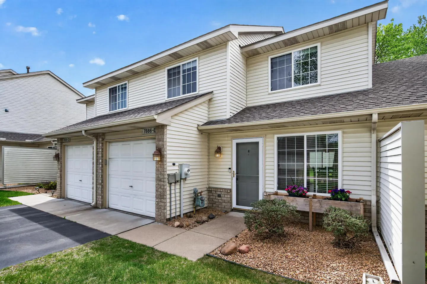 Beige two-story townhome with white garage doors, a gray front door, and a flower box under the window.
