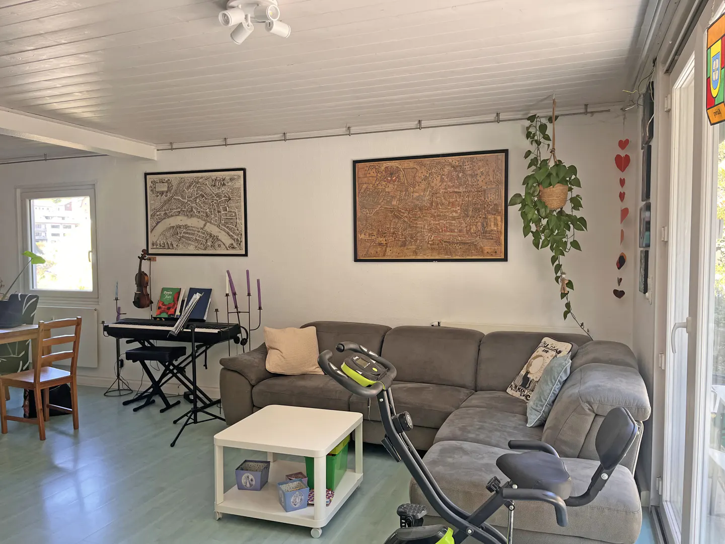 Living room with gray sectional sofa, exercise bike, keyboard, and framed maps on the wall. A hanging plant adds a touch of nature.