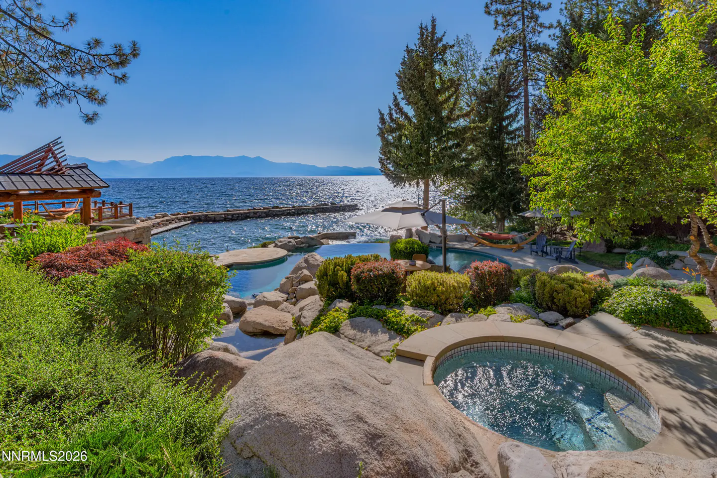 Lakefront property with a hot tub, pool, and gazebo. The water is blue and the sky is clear. There are trees and bushes around the property.