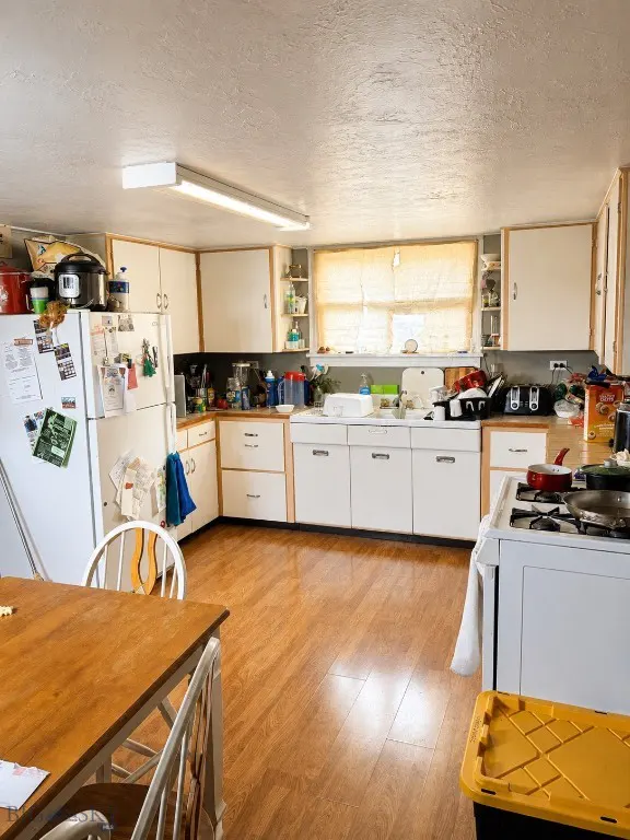 Kitchen with white cabinets, wood flooring, a white refrigerator, and a wooden dining table in the foreground.