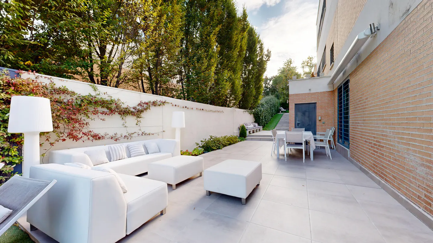 Outdoor patio with white furniture, including a sofa, ottomans, and dining set. A brick building and green trees are in the background.