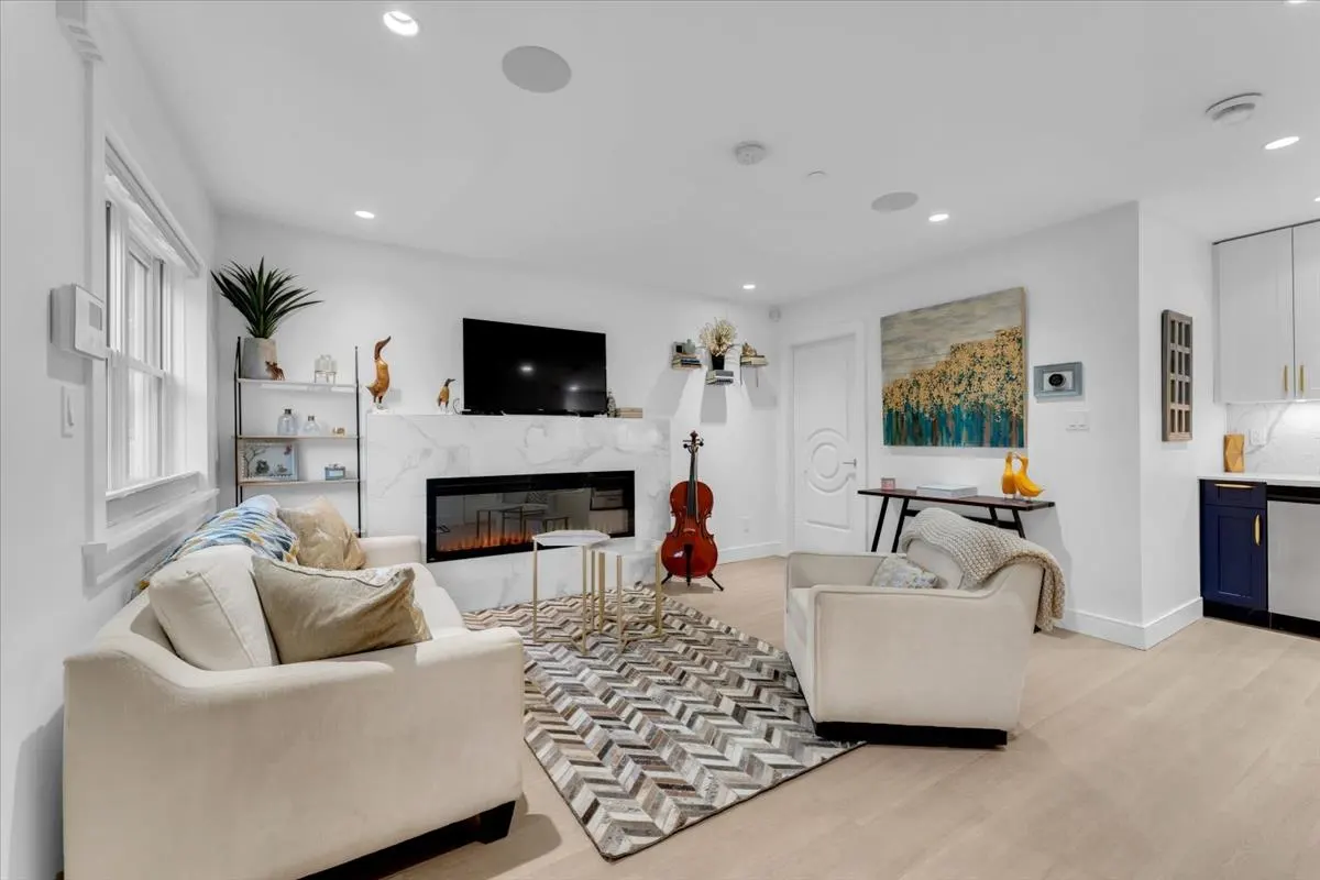 Bright living room with white sofas, a marble fireplace with a TV above, and a cello standing next to it. A patterned rug covers the floor.