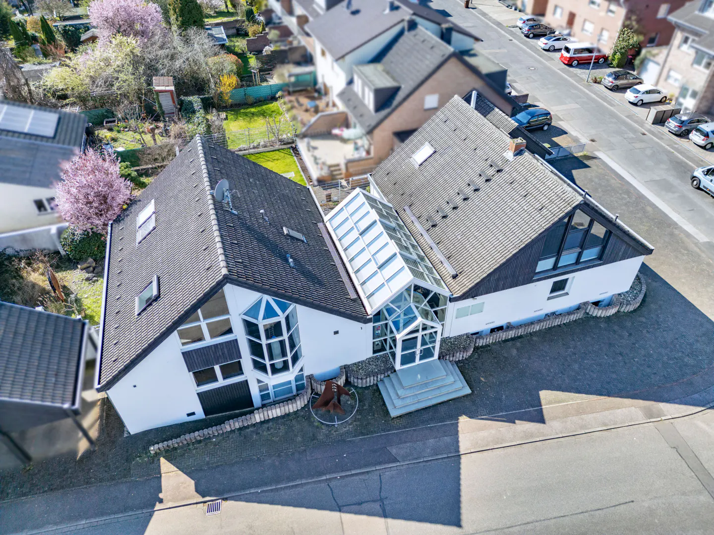 Aerial view of a white house with a glass conservatory connecting two sections, dark gray roof, and a sculpture in the front yard.