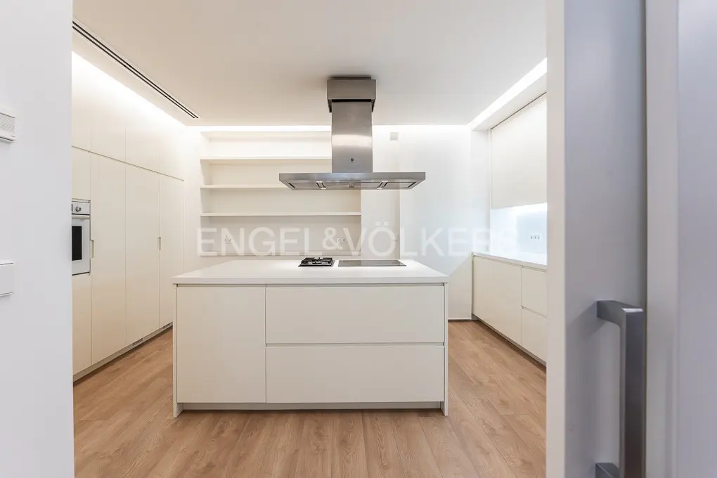 A modern, all-white kitchen with a central island, stainless steel range hood, and light wood floors.