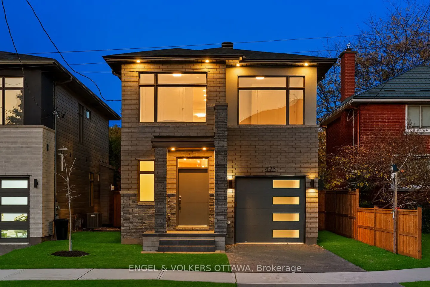 Two-story brick house with a gray door and garage. Lights illuminate the windows and exterior. Lawn and sidewalk in front.
