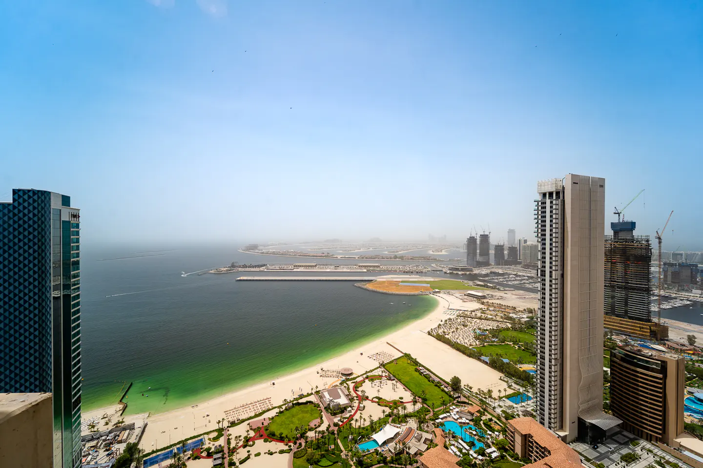 Aerial view of Dubai's coastline, featuring sandy beaches, turquoise water, and modern skyscrapers under a hazy blue sky.