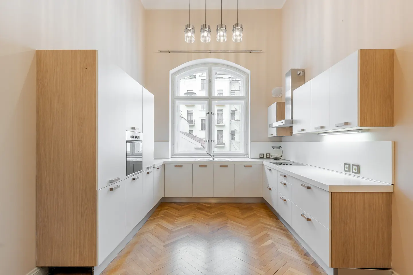 Bright kitchen with white cabinets, wood accents, and herringbone floor. A large arched window overlooks a city view. Modern pendant lights hang above.