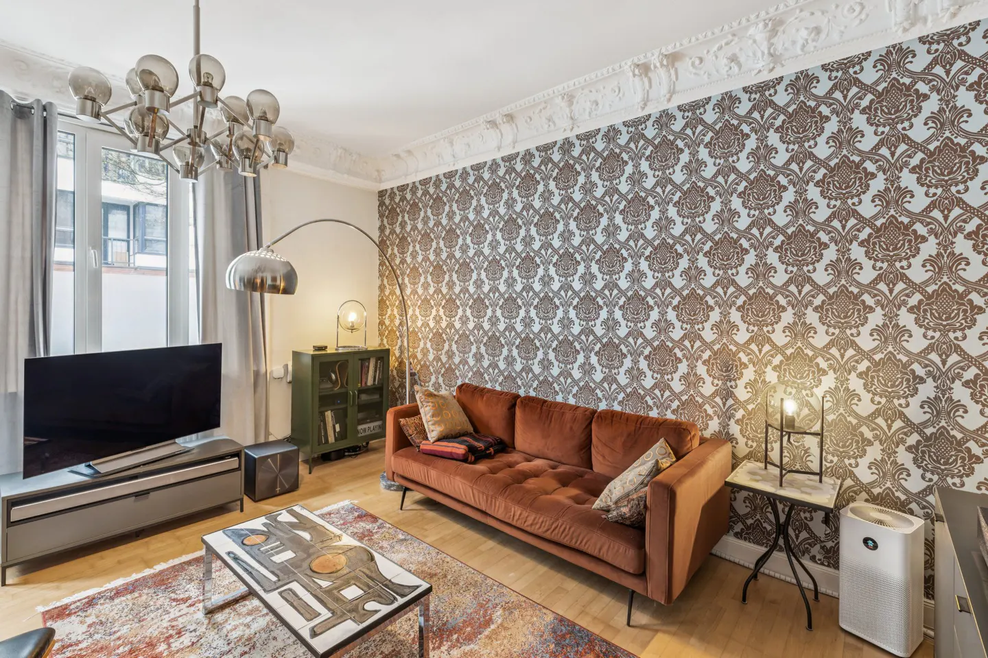 Living room with brown patterned wallpaper, a brown velvet sofa, and a modern chandelier.