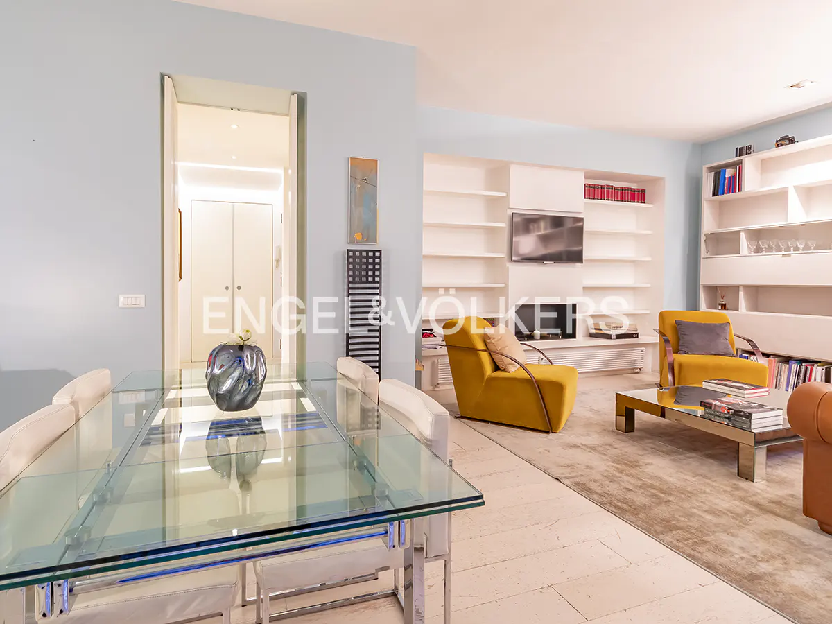 Bright living room with a glass dining table, yellow armchairs, and white bookshelves against light blue walls.