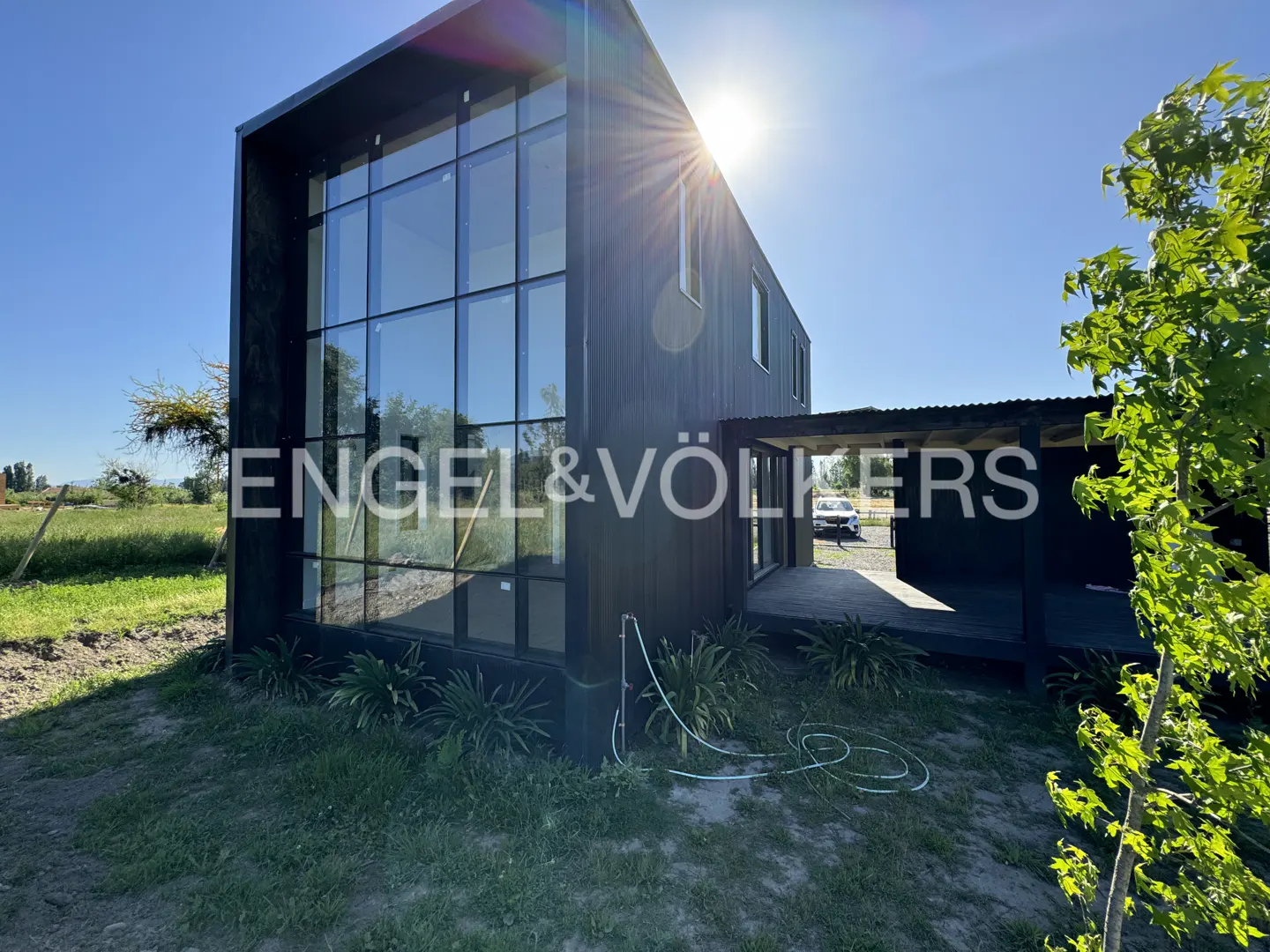Modern black house with floor-to-ceiling windows, a covered patio, and a sunny yard under a clear blue sky.