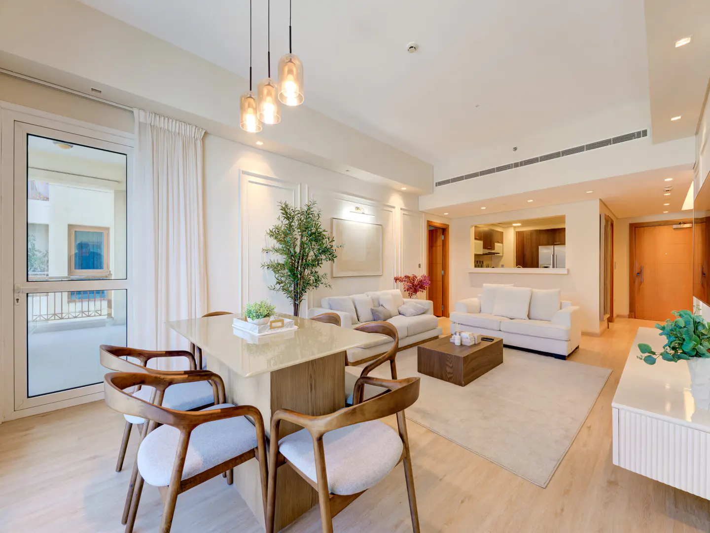 Bright, open-plan living and dining area with white sofas, wood table and chairs, and pendant lights. Kitchen visible in the background.