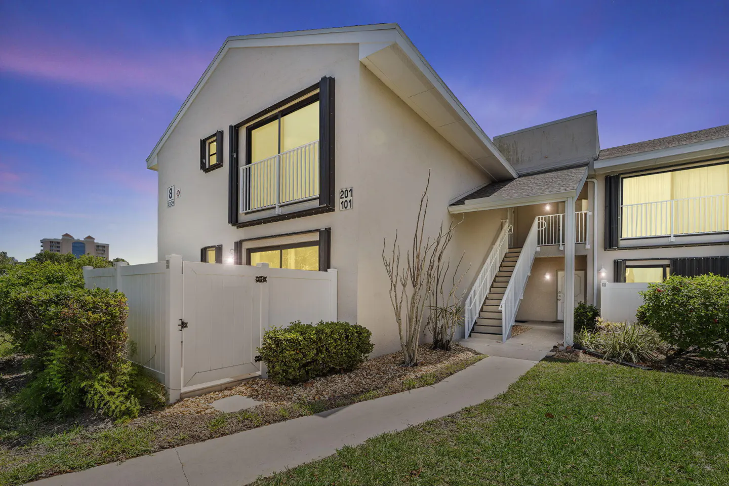 Exterior view of a two-story condo with white walls, black trim, and a white fence. A walkway leads to stairs.