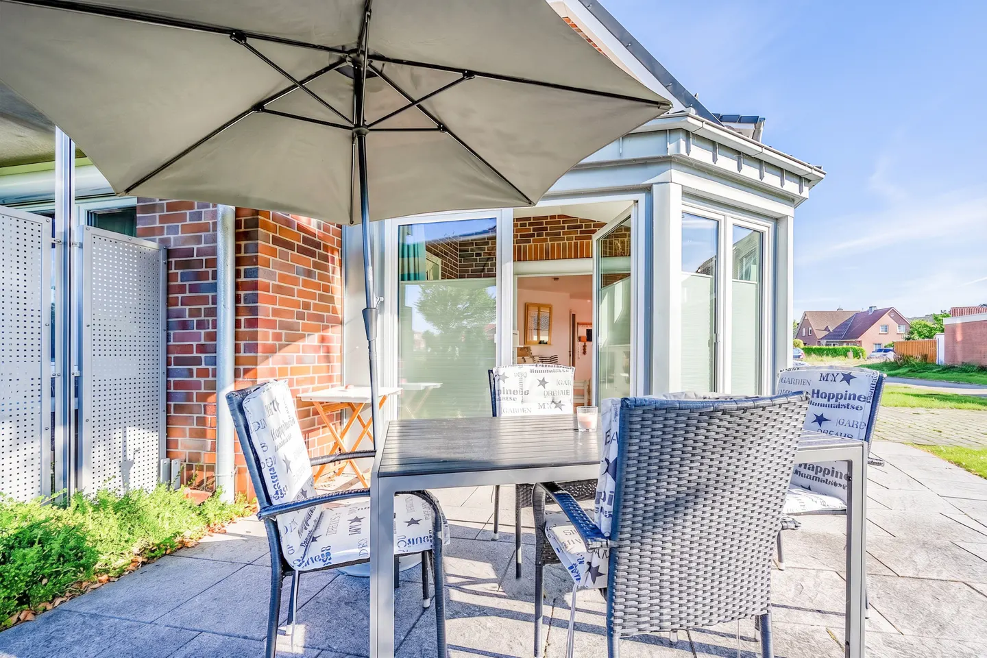 Outdoor patio with a gray table, four chairs, and a large umbrella. A brick building and a sunroom are in the background.