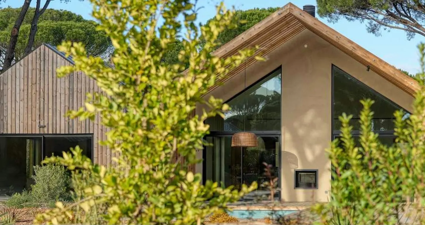Modern home exterior with a tan stucco A-frame and a wood-sided building, partially obscured by green foliage.