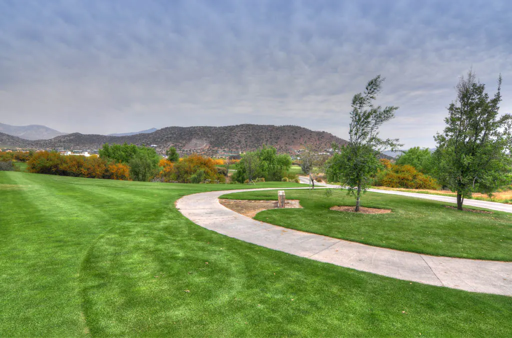 A winding concrete path cuts through a lush green golf course, with trees, mountains, and a cloudy sky in the background.