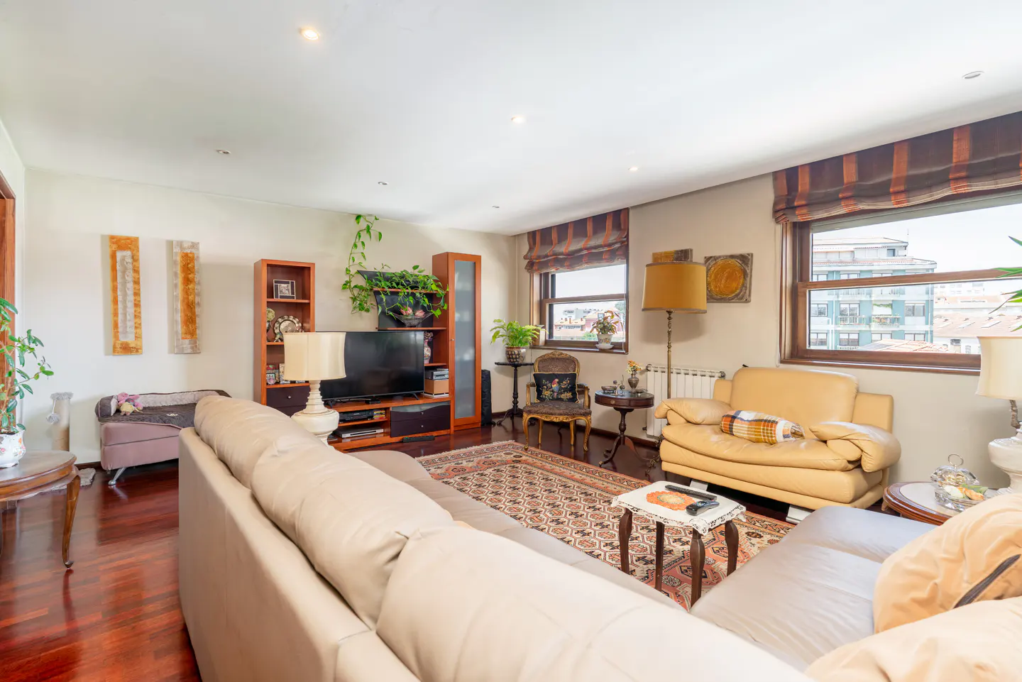 Living room with a beige sofa, patterned rug, wooden floors, TV unit, and large windows with orange-striped blinds.