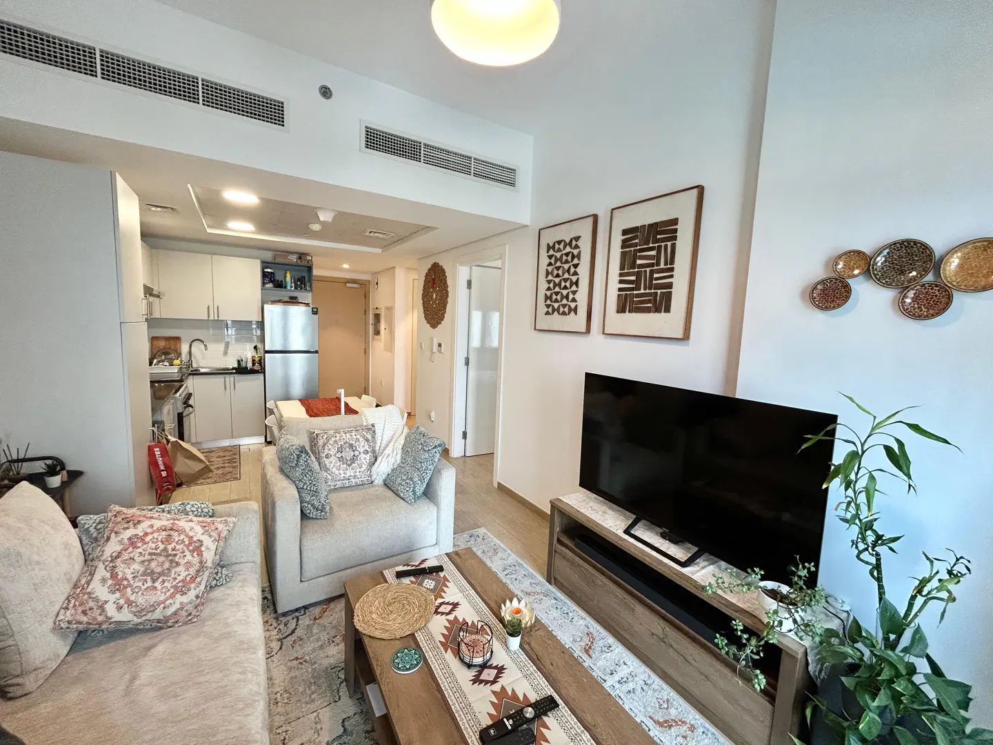 A bright, modern apartment living room with a sofa, armchair, TV, and open kitchen. Artwork adorns the white walls.