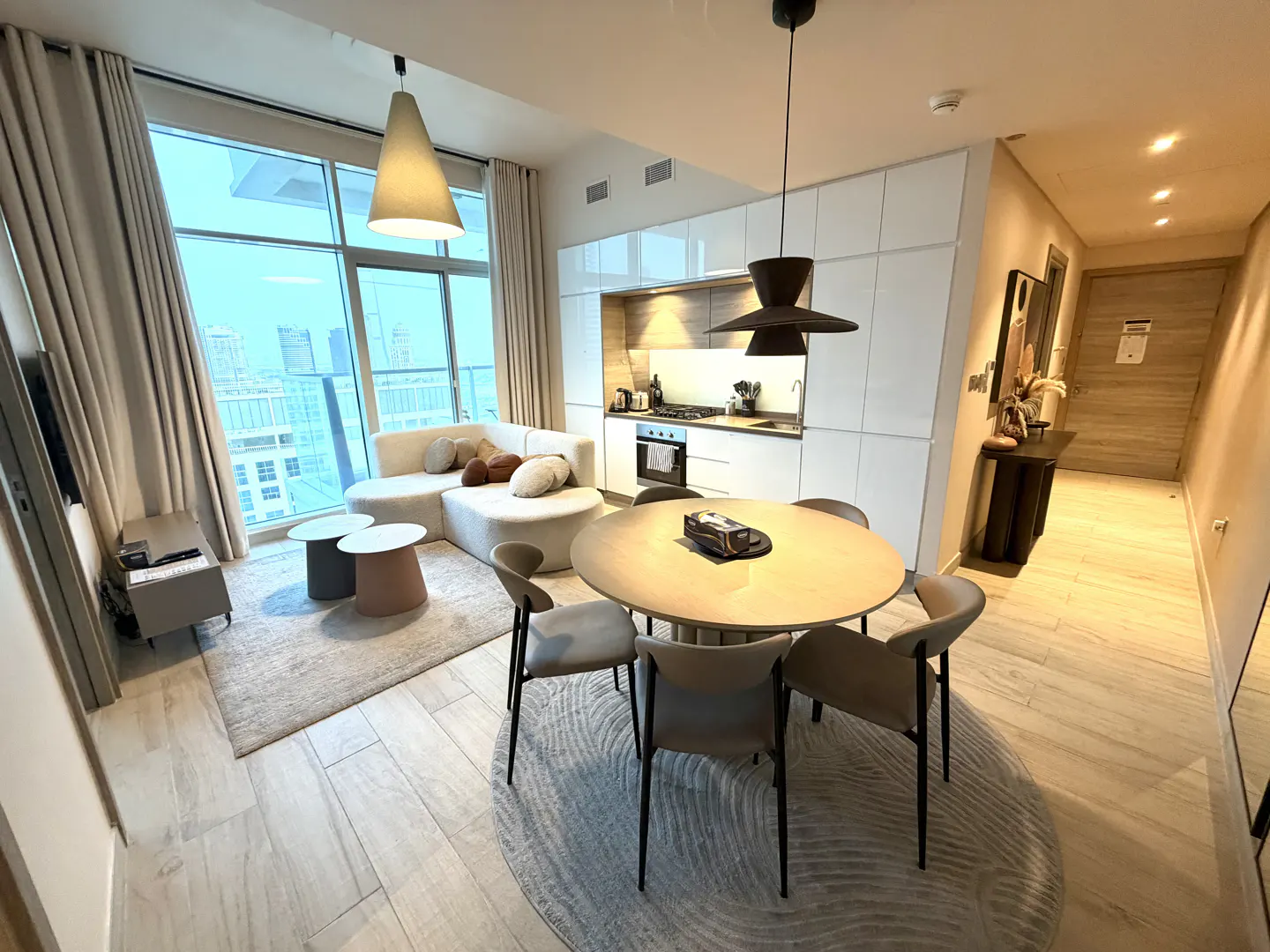 A modern apartment interior with a round dining table, a white sofa, and a kitchen. Large windows offer a city view.