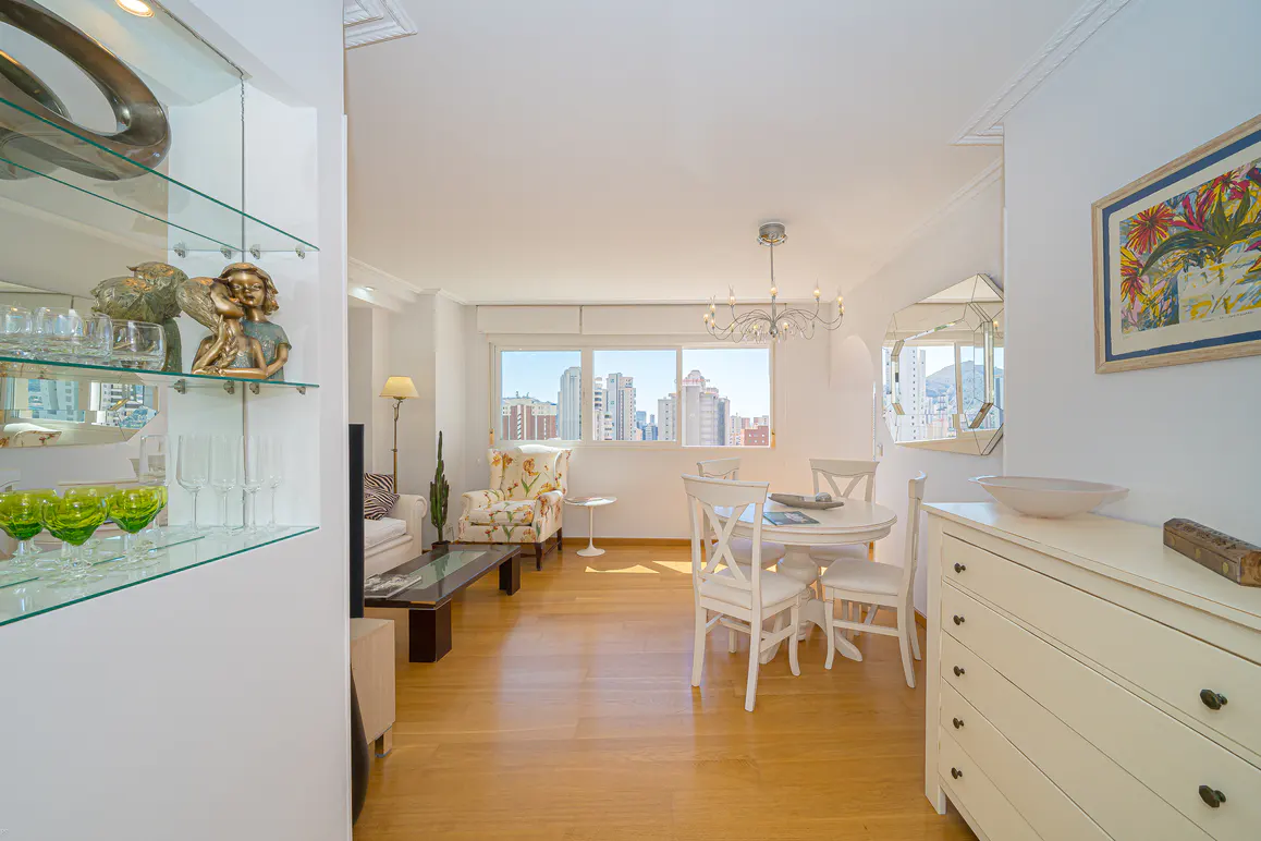 Bright living room with hardwood floors, white walls, and city views. A glass shelf displays decorative items. A white dining set sits near a window.