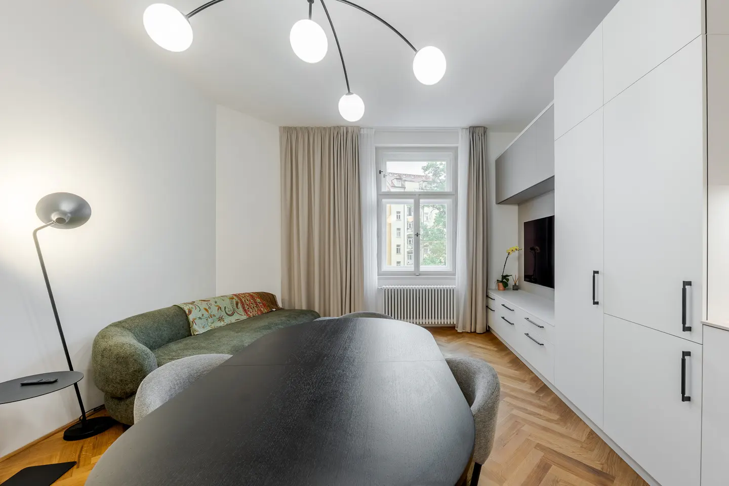 Modern living room with a black oval dining table, green sofa, white cabinets, herringbone wood floors, and globe lighting.