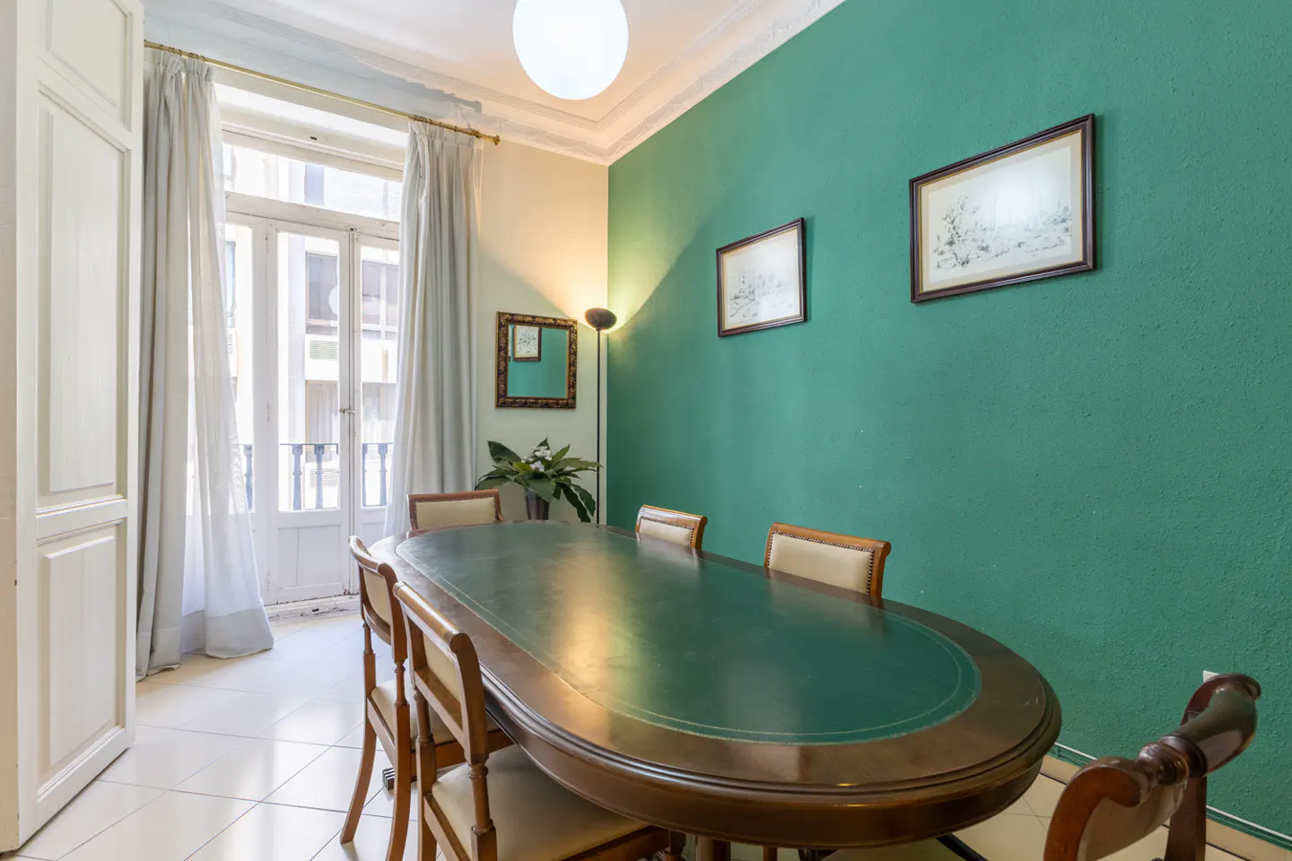 A dining room with a green wall, a large oval table, and wooden chairs. A window with white curtains is on the left.